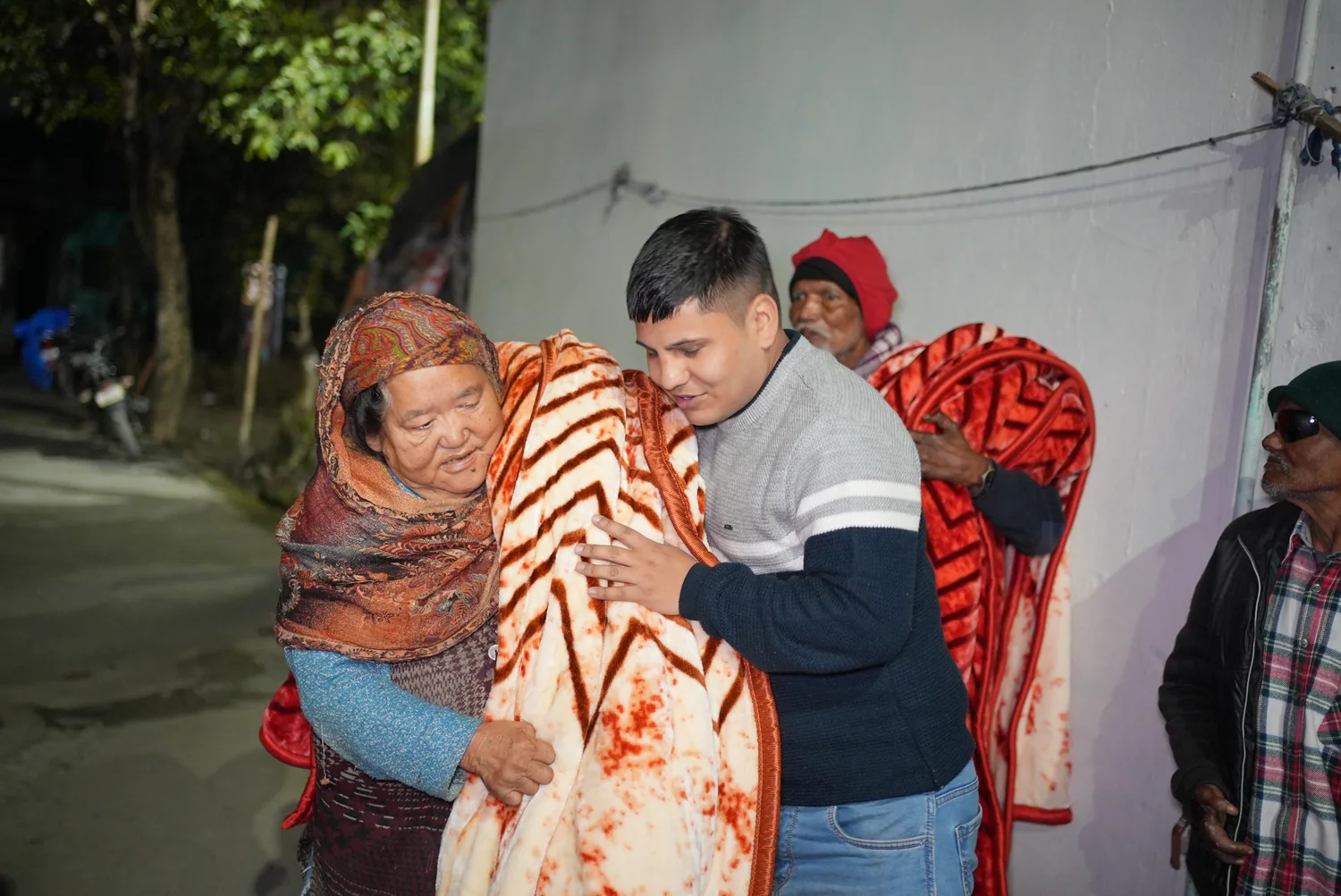 A team member helps an older woman wrap a warm patterned blanket around her shoulders.