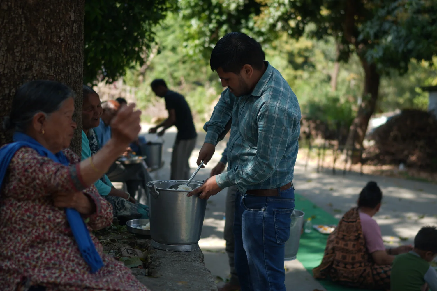 A team member carries prepared food to older residents seated outdoors.