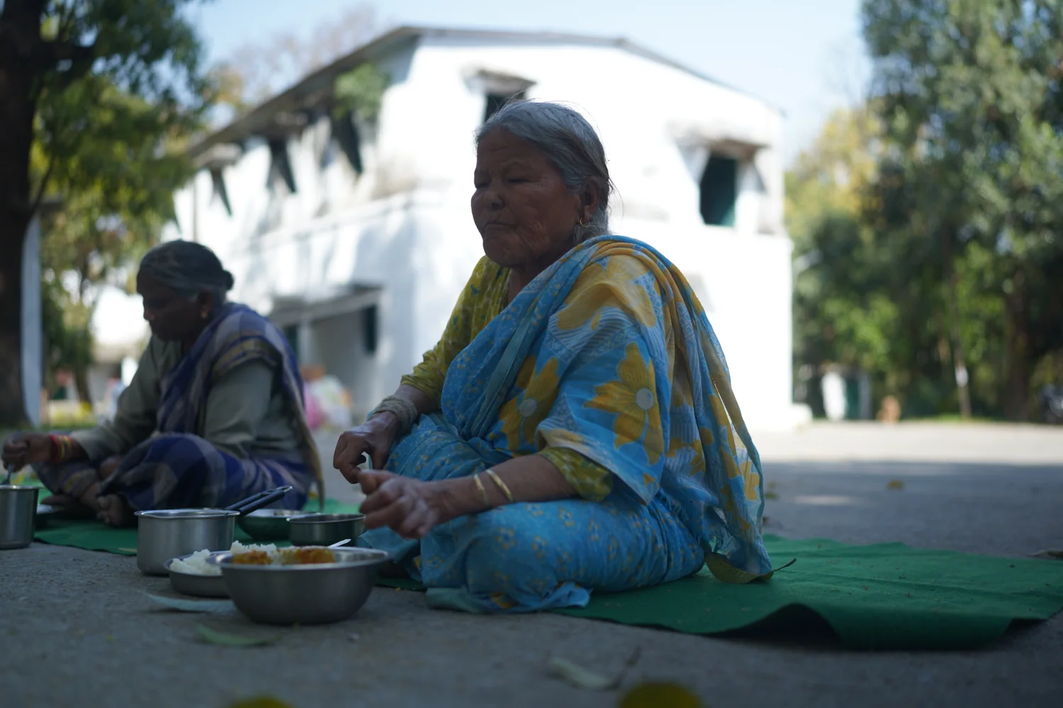 An older woman sits with her meal in the shaded gathering area.