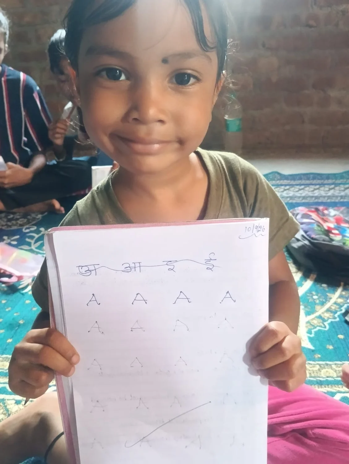 A young learner shows a page with Hindi letters and repeated English letter A practice.