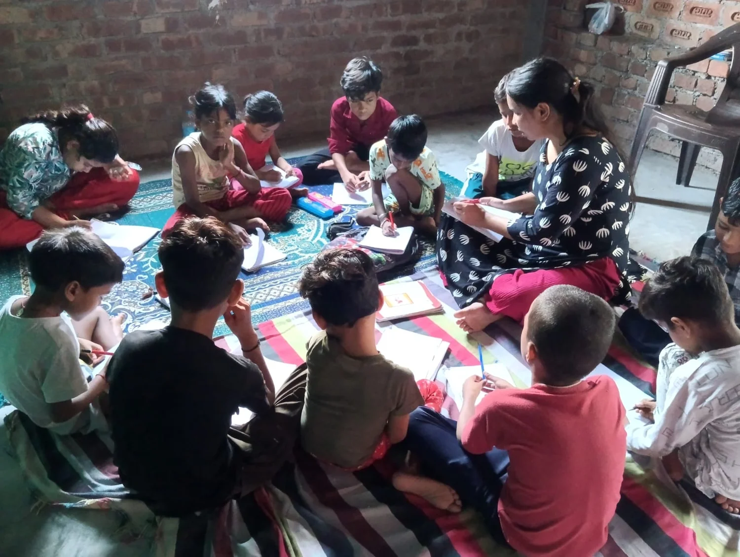 Children sit with a teacher around open notebooks during a group study session.