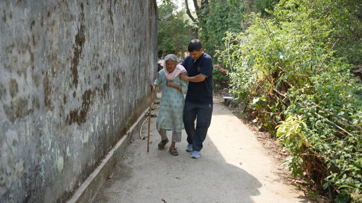 A volunteer assisting an older resident along a narrow lane