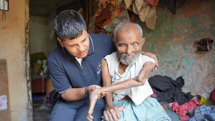 A volunteer supporting an older man seated inside his home