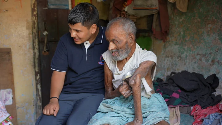 A Dev Bhoomi Samiti volunteer sitting beside an older man during a home visit